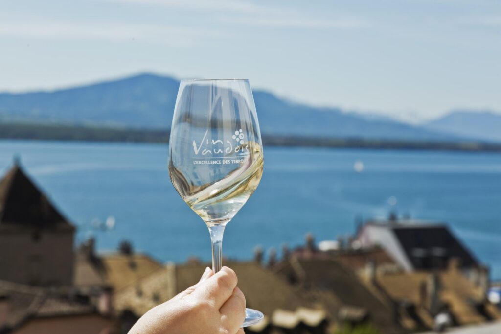 Verre de vin blanc levé face au Léman, au-dessus des toits de Lavaux